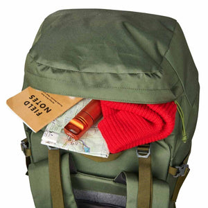Dark green 65L backpack by Kelty, top opening reveals a 'FIELD NOTES' notebook, map, orange flashlight, and red knit hat.