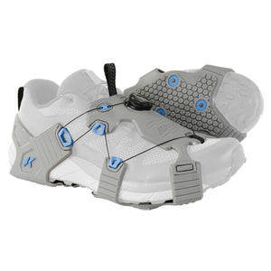 Grey ice traction device by Korkers with blue eyelets, a black dial lacing system, and a honeycomb-patterned sole, on a white sneaker.