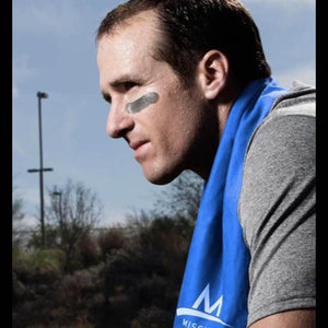 Blue cooling towel by Mission with a visible M logo, draped around a man wearing a grey t-shirt and eye black.