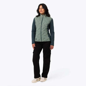 Women's sage green and teal hybrid heated jacket by Mobile Warming with quilted diamond pattern, worn with black cargo pants and beige sneakers.