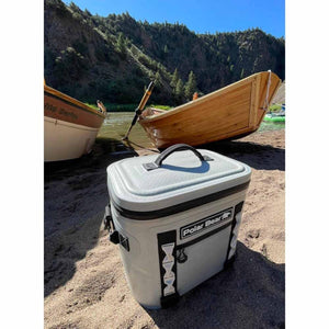 Grey cooler bag by Polar Bear Coolers, soft-sided with a top handle, on sand with boats and a river.