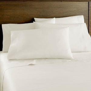 Cream sateen sheet set by Shavel on a neatly made bed with four matching pillows against a wooden headboard.