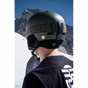 Dark green ski helmet by Smith Optics with a yellow Mips logo and brown goggle strap, worn from the back in a snowy mountain landscape.