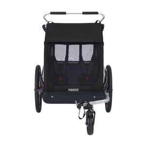 Black 2-seat bike trailer by Thule with mesh windows, two harnesses, large side wheels, and a front swivel wheel.