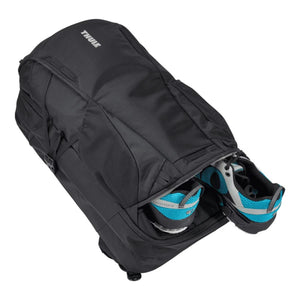 Black laptop backpack by Thule with blue and grey running shoes in a bottom side compartment, angled diagonally.