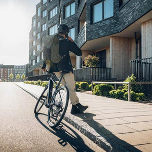 Olive green commute backpack by Thule worn on a person standing with a bicycle in an urban setting.