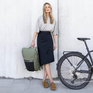 Olive green and black commuter backpack by Thule, held by a person standing next to a bicycle.