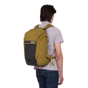 Olive green and black backpack by Thule, featuring a top flap, black lower panel with logo, and a side pocket, worn on a person's back.