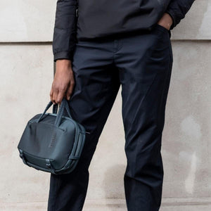 Dark teal crossbody bag by Thule, held by a person in a black jacket and dark pants.