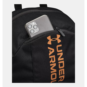 Black laptop backpack by Under Armour with an orange logo and a smartphone in a zippered front pocket, cropped view.