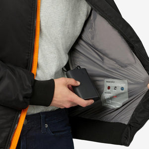 Black Battery Heated Bomber Jacket by Venture Heat with orange zipper, grey interior lining, and a hand inserting a black power bank into an internal pocket.