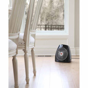 Black whole room heater by Vornado with a white spiral grill, on a light wood floor by a window.