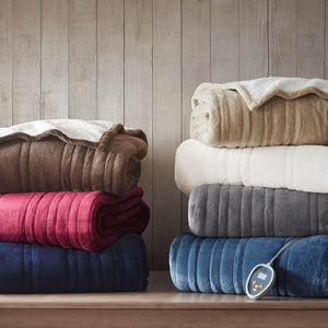 Multiple ribbed heated plush to berber blankets by Woolrich in various colors, with a controller visible, stacked on a wooden surface.