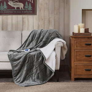 Gray and white reversible heated throw by Woolrich, with a quilted plush side and controller, draped on a sofa.